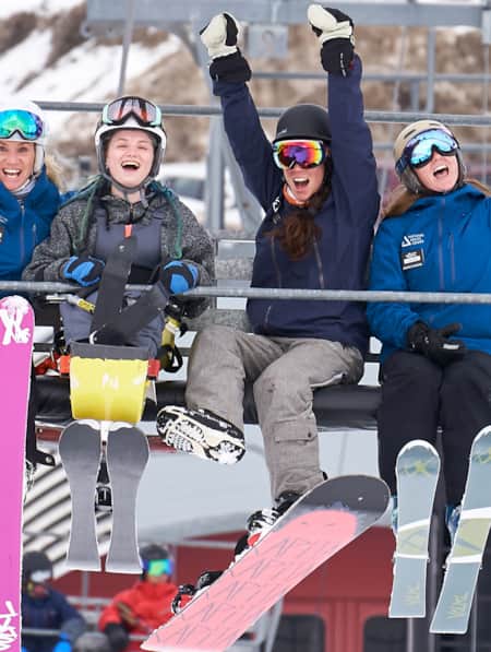 Four women on a ski lift, dressed in winter clothes and ski gear, are smiling and excitedly throwing their hands up in the air.