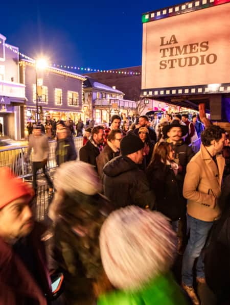 A crowd of people standing outside on the side of a street in cold weather, bundled up in winter clothing.