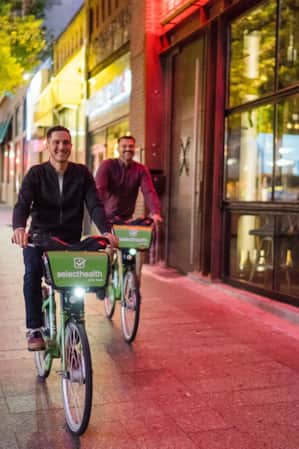 Two men riding city bikes on a sidewalk in the evening, with city buildings in the background.