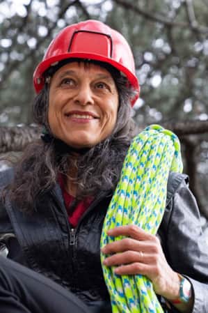 A woman sitting on a tree branch, wearing a red helmet and carrying a neon green rope.