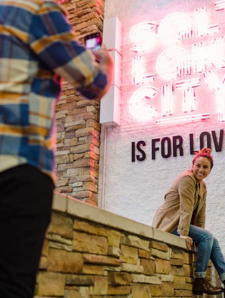 A woman sitting and smiling at the base of a sign that reads 'Salt Lake City is for lovers,' while having her picture taken.