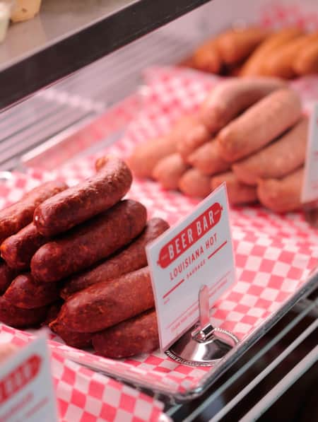A deli case displaying various types of sausage links on silver trays lined with red and white checkered paper.