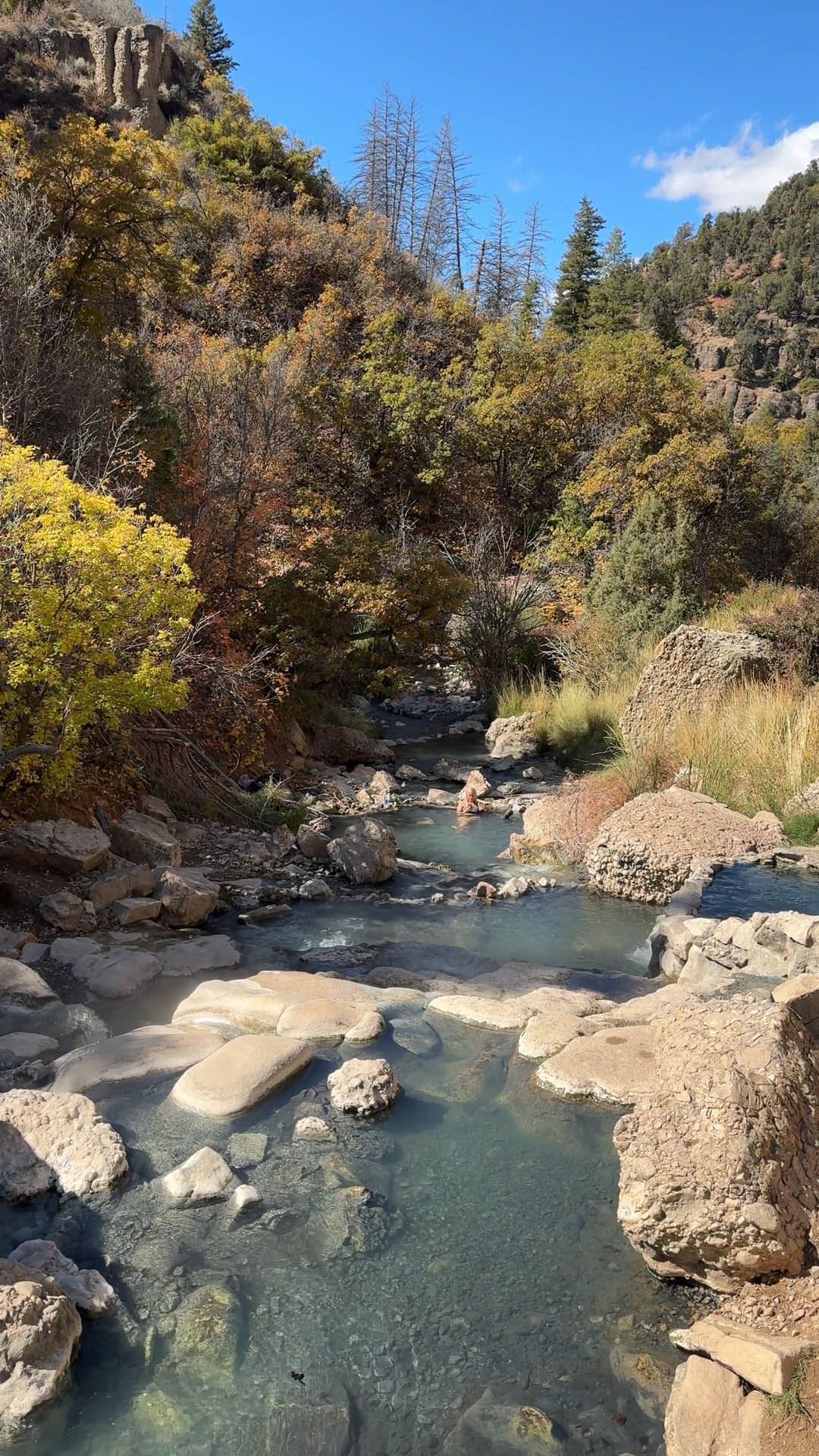 Rocky cliffs envelop the blue hot springs fed by bubbling waterfalls along a creek.