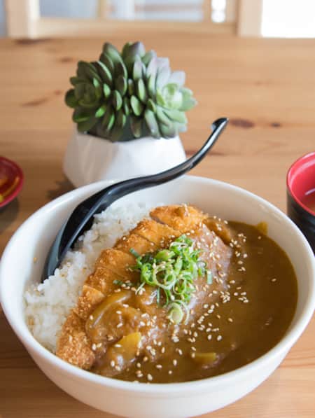 A bowl of rice topped with thick brown curry sauce, placed on a wooden table in a restaurant.