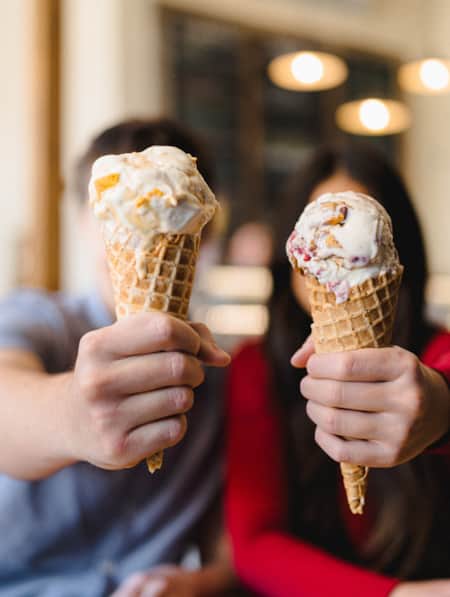 Two people out of focus, each holding up ice cream cones that are in focus, covering their faces.