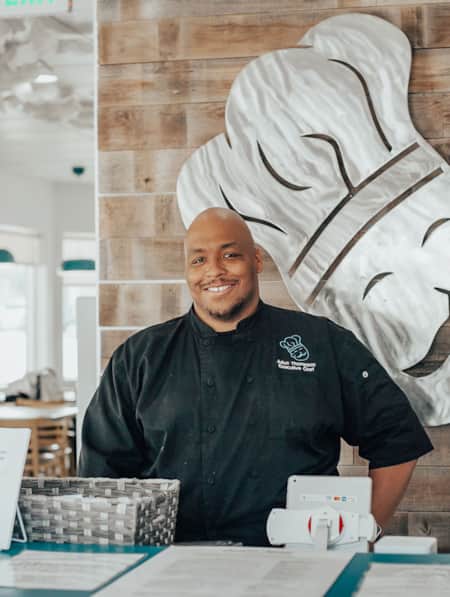 A man wearing a chef's uniform, smiling behind the counter at a restaurant.