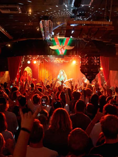 A crowded room with red lights, where people are looking at and listening to a live band in an ornate setting.