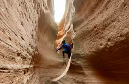 A person carefully climbing through a narrow, winding slot canyon with steep rock walls on either side.