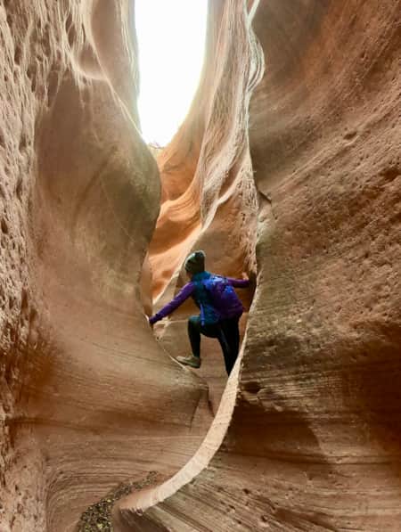 A person carefully climbing through a narrow, winding slot canyon with steep rock walls on either side.
