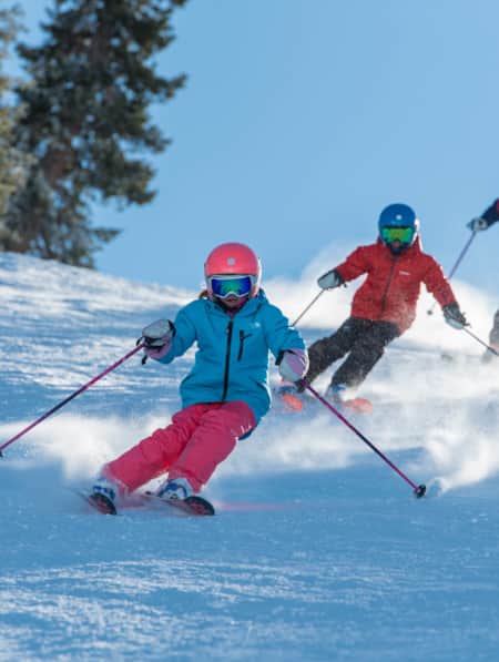 Two young children and an adult skiing down a run, with pine trees on each side.