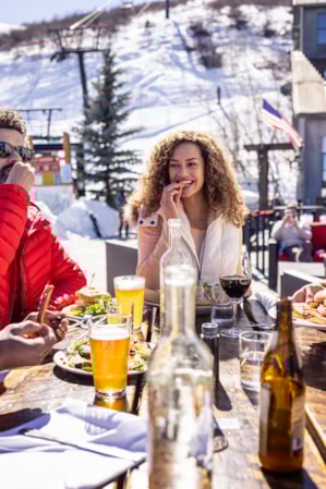 People at a ski resort, bundled up in winter clothing, eating and drinking at a wooden table outside.