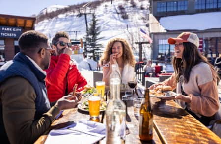 People at a ski resort, bundled up in winter clothing, eating and drinking at a wooden table outside.