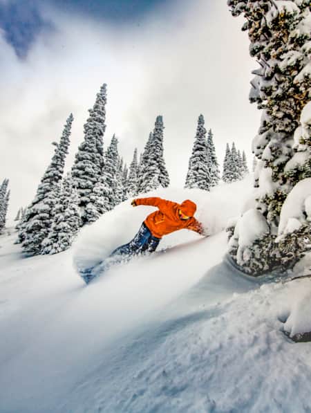 A snowboarder coming down the mountain on his edge, kicking up a lot of snow.