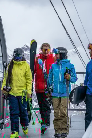 Four skiers and snowboarders, dressed warmly, are holding their equipment while talking and walking off a gondola.
