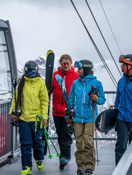 Four skiers and snowboarders, dressed warmly, are holding their equipment while talking and walking off a gondola.