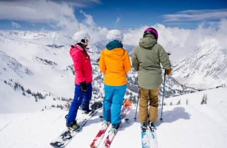 A backside view of three skiers at the top of a mountain, overlooking a canyon of snow-covered peaks.