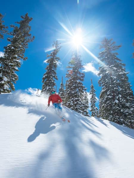 A man in a red winter jacket skiing down a mountain slope on a clear blue sky day.