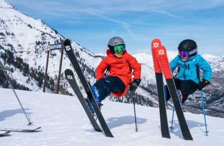 Two kids in ski gear on top of a mountain, propping themselves up with their skis and poles.