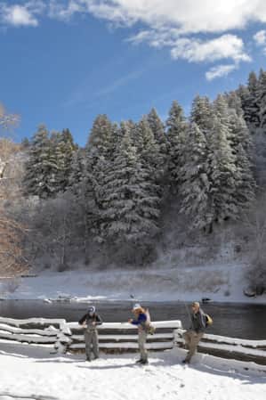 Three people preparing to fly fish by a river in a snowy landscape.