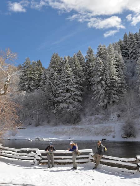 Three people preparing to fly fish by a river in a snowy landscape.