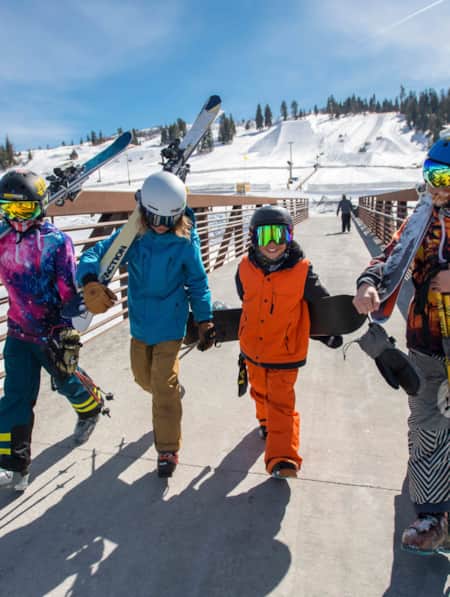 Four kids walking side by side, carrying skis and snowboards as they cross a bridge, with ski slopes in the background.