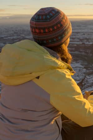 A woman in winter clothing looking into the distance at a setting sun over a canyon dusted with snow.