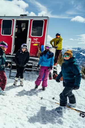 A group of people geared up to ski at the top of a mountain, being dropped off by a snowcat.