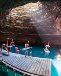 Four women on paddle boards in water doing yoga poses, following an instructor on a wooden dock, all within an underground crater.
