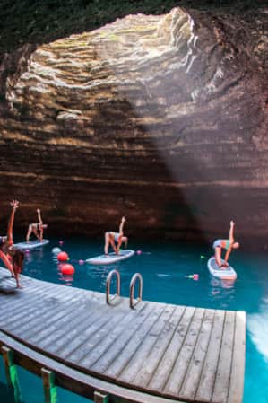 Four women on paddle boards in water doing yoga poses, following an instructor on a wooden dock, all within an underground crater.