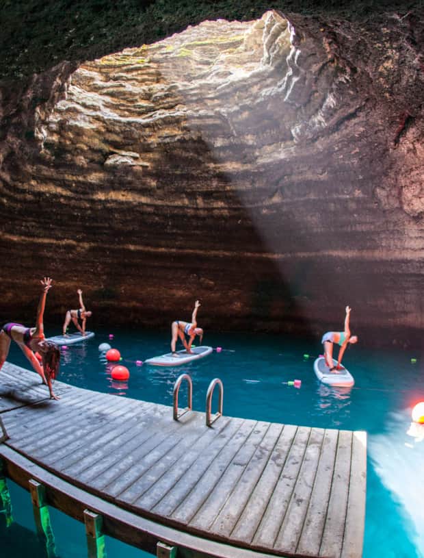 Four women on paddle boards in water doing yoga poses, following an instructor on a wooden dock, all within an underground crater.