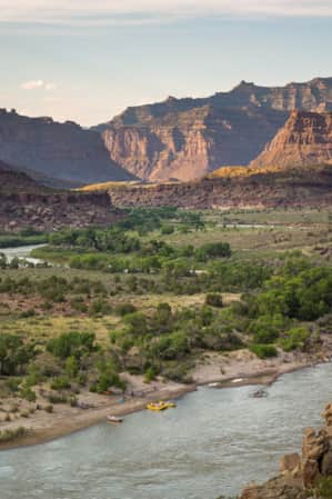 A wide river winding through a rocky canyon of buttes, with some rafters resting at the shore.
