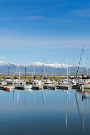 Multiple sailing boats docked on a lake, with snow-capped mountains in the background.