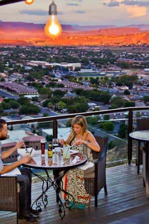 A man and a woman enjoying a meal at sunset on a restaurant patio overlooking the city.