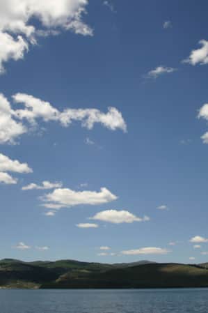 An empty lake with green hills along the shore and a clear blue sky with a few clouds.