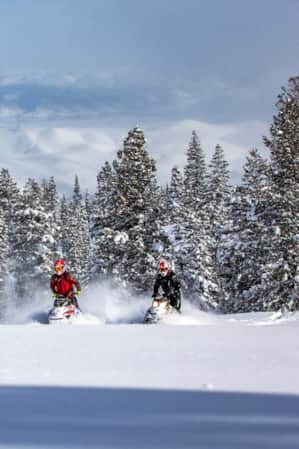 Two snowmobilers driving through the snow in the mountains, with a large lake and towering mountains in the backdrop.