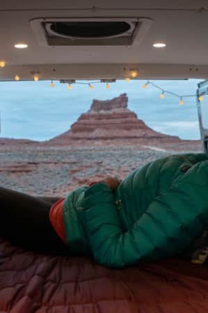 A woman warmly dressed, sitting in a van with a bed set up in the back, gazing out at a large rock formation in the desert.