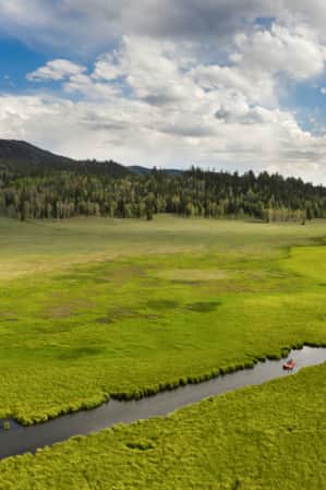 A narrow stream winding through a grassy field, with people kayaking on it and green hills covered with trees on each side.