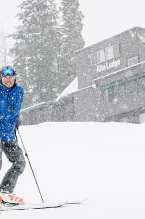 A skier smiles in front of a ski lodge while it is snowing.