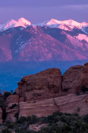 Purple and blue-hued mountains at sunset with snow-capped peaks, and red rock formations prominently featured in the foreground.