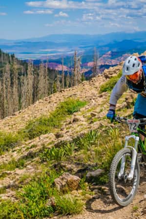 A man mountain biking on a dirt path along the side of a mountain, with additional mountain ranges in the backdrop.