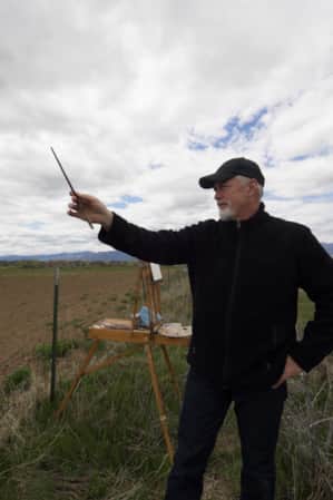 A man in an open field with an easel, holding up his paintbrush.