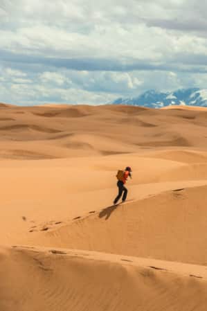 woman hiking through sand dunes with snow-capped mountains in the background.