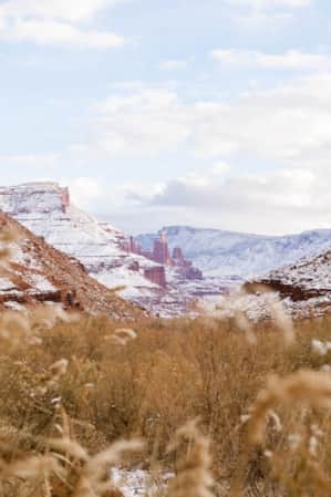 A valley filled with hay lightly dusted in snow, nestled at the base of large red rocky mountains with flat plateaus at the top and hoodoos in the background.
