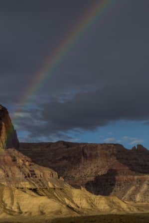 Half of a rainbow arcs over a desert landscape with tall monument rock formations.
