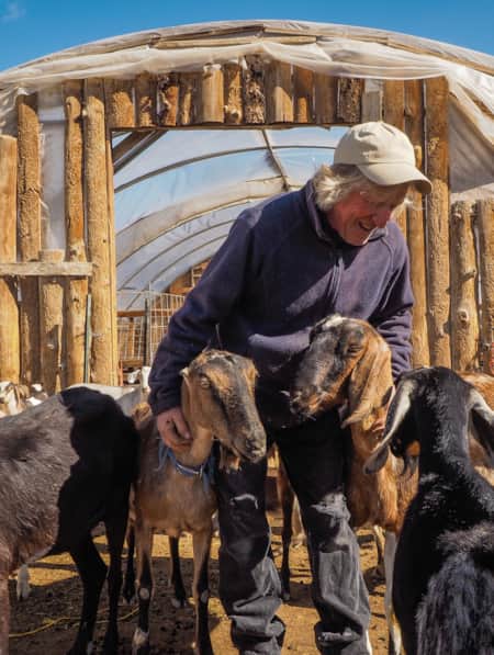 A man standing in front of a greenhouse framed in logs, looking down and hugging a group of goats.