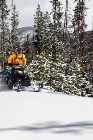 Two people driving snowmobiles on a snowy path through the mountains on a cloudy day.
