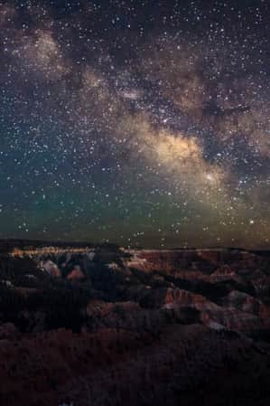 An expansive view of layered rock mountains with trees growing on them, under a starry night sky.