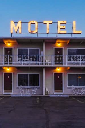 The front view of a motel with a parking lot in front, where each room door is illuminated by a porch light.