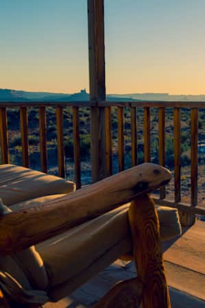 A wooden bench with fabric cushions on a porch, overlooking an open field in the desert.