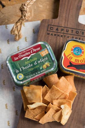 A wooden serving board with two tins of canned fish and crispy pita chips on top.